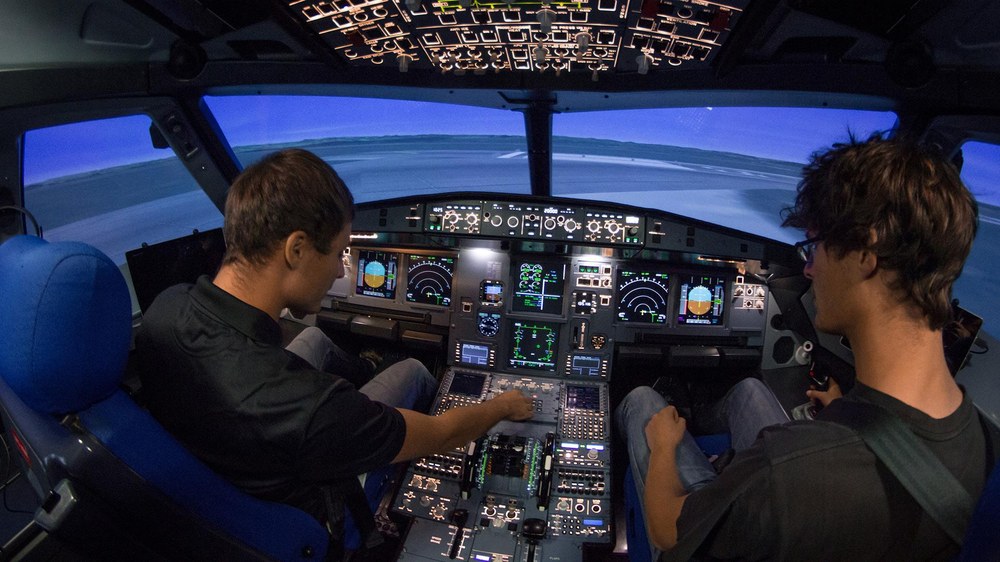 Two people seated in an airplane cockpit simulator with illuminated control panels, focusing on flight instruments. The scene conveys concentration and teamwork.