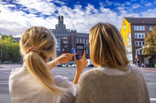 Zwei Frauen stehen vor einem Gebäude mit einer U-förmigen auf dem Dach und schauen gemeinsam auf das Display eines Smartphones.