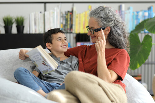 Eine ältere Frau mit Brille mit integrierter digitaler Kamera auf einer Couch neben einem Jungen und ein Buch in der Hand haltend.