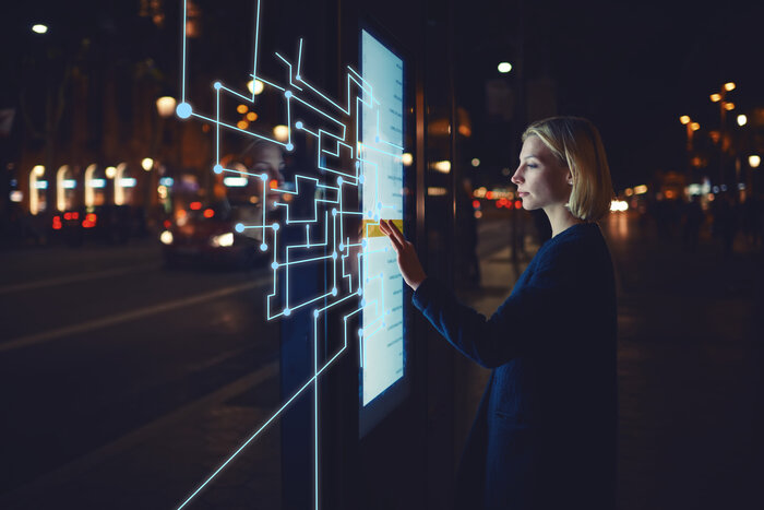 Ein Frau bedient einen beleuchteten digitalen Touchscreen mit grafischen Linien und Punkten als Interface-Overlay in einer Stadt bei Nacht.