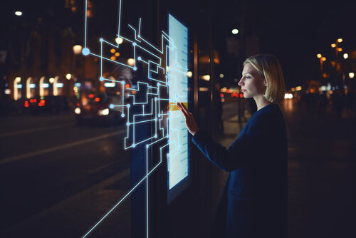 Ein Frau bedient einen beleuchteten digitalen Touchscreen mit grafischen Linien und Punkten als Interface-Overlay in einer Stadt bei Nacht.