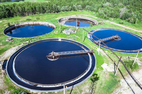 Vier große runde Klärbecken einer Abwasserreinigungsanlage, umgeben von grüner Vegetation und Bäumen.