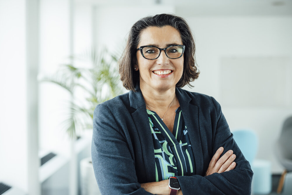 A portrait photo of a woman in an office space. She has her arms crossed and is wearing a blouse with various patterns and a dark-coloured blazer.