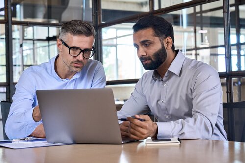 Zwei Männer in Hemden sitzen an einem Tisch und schauen gemeinsam auf das Display eines Laptops.
