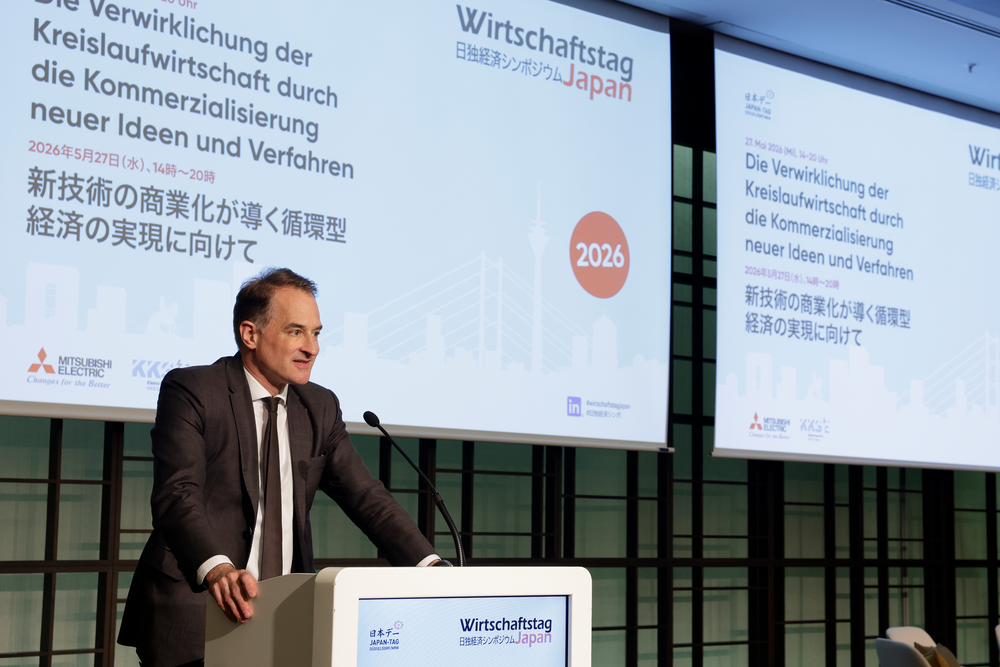 A man speaks at a podium during the Wirtschaftstag Japan conference. The presentation screen features text in German and Japanese, with a 2026 logo.