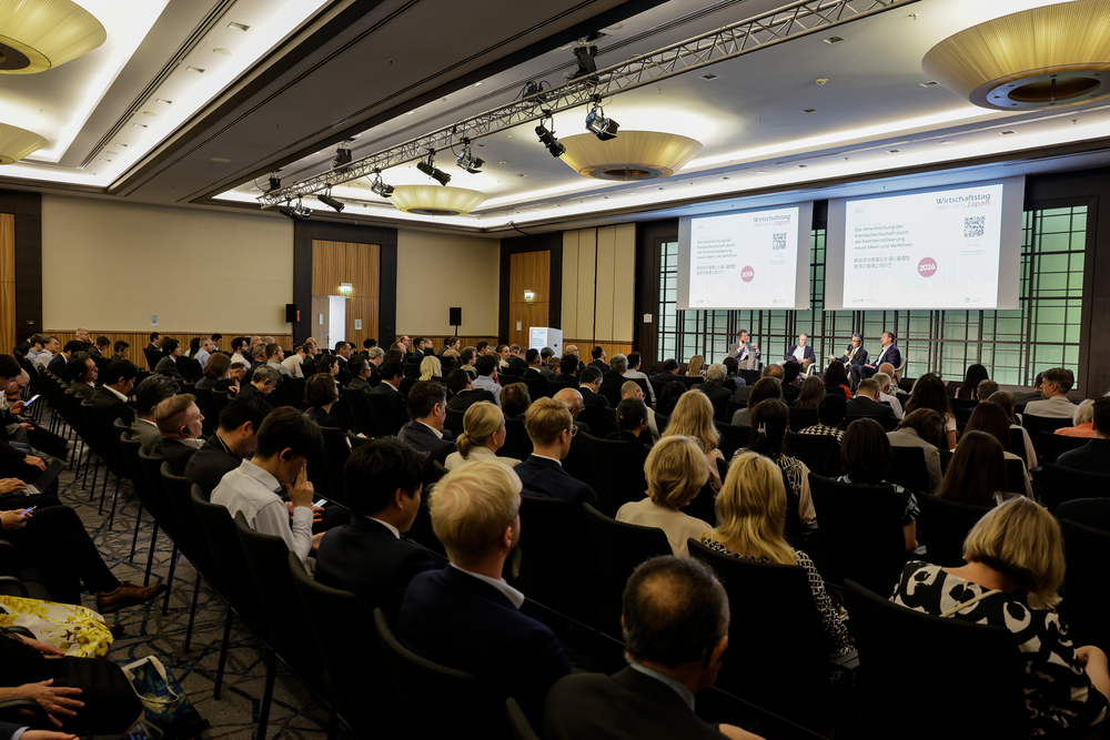 A large conference room filled with people seated in rows facing a panel of four speakers on stage. Two large screens display presentation slides. The atmosphere is professional and focused.