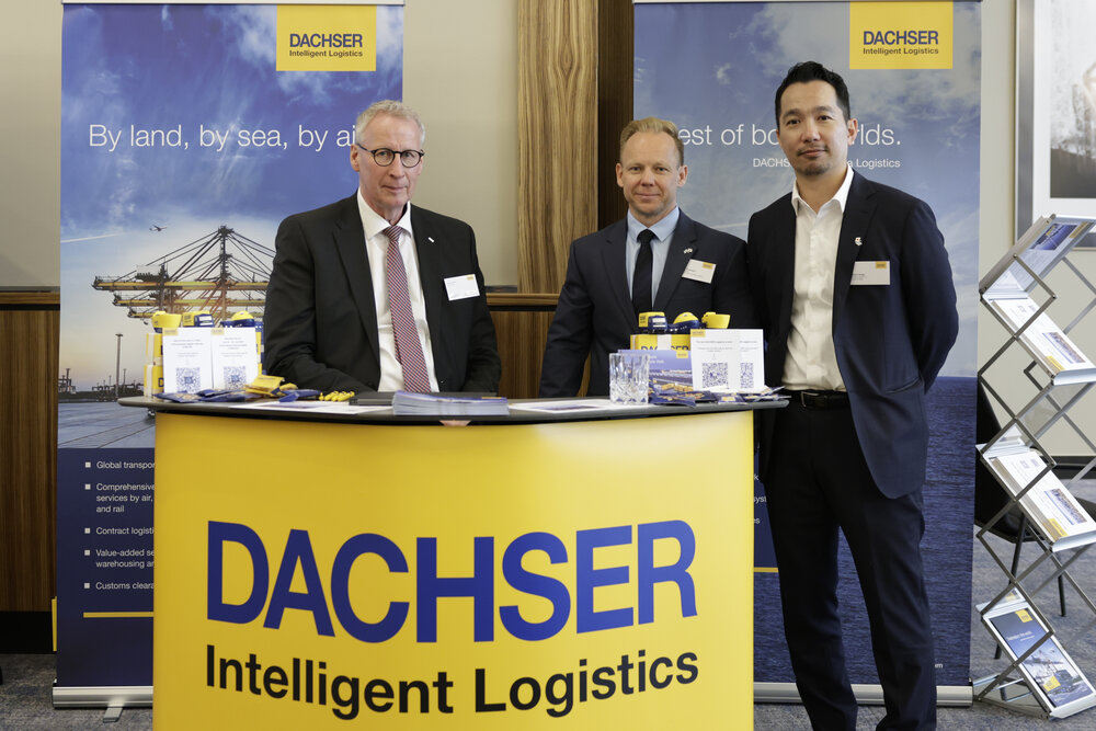 Drei Männer in Anzügen stehen hinter einem gelben Messestand mit Aufschrift Dachser Intelligent Logistics, im Hintergrund sind Banner aufgestellt.