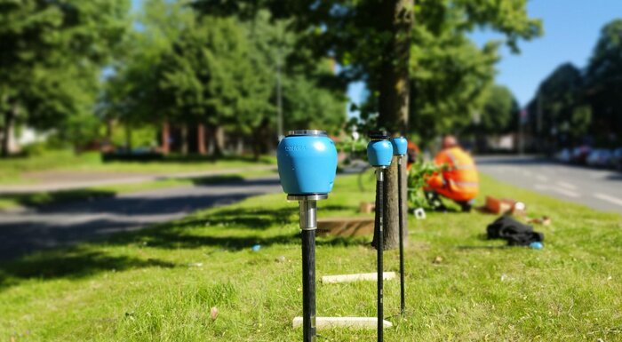 Mehrere blaue Messgeräte auf schwarzen Stangen im Gras am Straßenrand, im Hintergrund zwei Menschen in orangefarbener Arbeitskleidung.