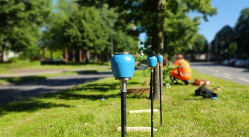 Mehrere blaue Messgeräte auf schwarzen Stangen im Gras am Straßenrand, im Hintergrund zwei Menschen in orangefarbener Arbeitskleidung.