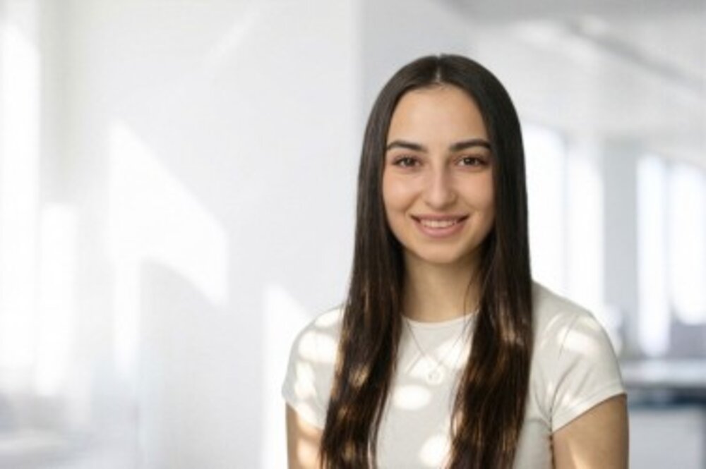 A portrait photograph of a woman with long straight hair wearing a white t-shirt against a bright background.