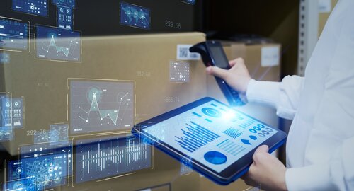 Eine Person scannt Kartons mit einem Handscanner und hält ein Tablet mit digitalen Diagrammen und Analyseanzeigen in der Hand.