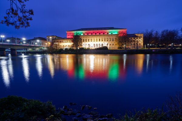 Ein großes historisches Gebäude am Flussufer bei Nacht, das in den Farben Grün, Weiß und Rot angestrahlt wird, und sich im Wasser spiegelt.