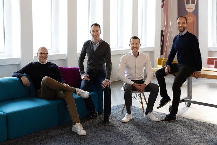 Vier Männer sitzen und stehen in einem modernen Büro, vor großen Fenstern und auf einer Couch, einem Stuhl und einem Rolltisch.