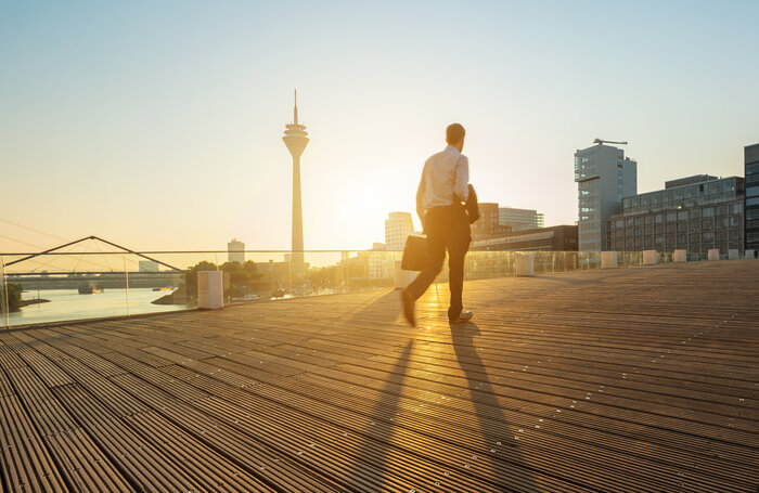 Ein Mann mit Aktentasche geht bei Sonnenuntergang über eine Holzterrasse in Düsseldorf, im Hintergrund der Rheinturm.