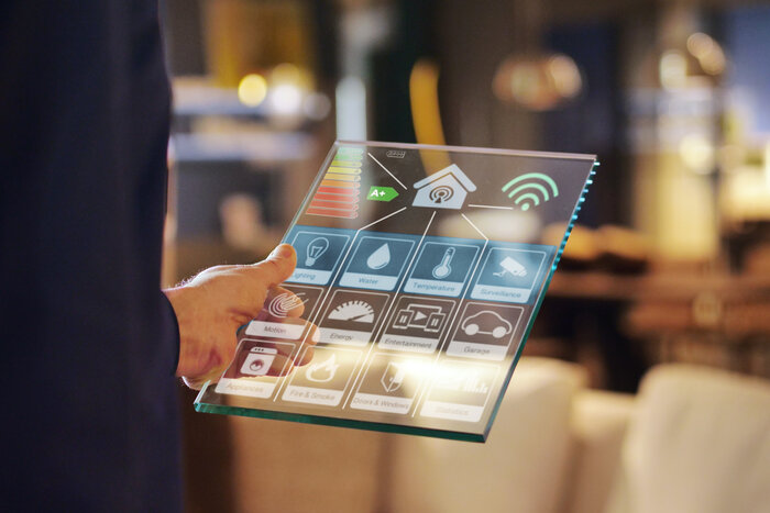 Eine Hand hält ein transparentes Tablet mit Smart-Home-Steuerung, unter anderem mit Symbolen für Beleuchtung, Wasser, Energie und Sicherheit.