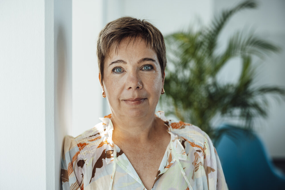 A portrait photograph of a woman with short hair wearing a patterned blouse standing indoors near a white wall with a plant in the background.