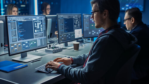 Mehrere Personen arbeiten in einem Büro an Computern mit großen Monitoren, die Programmcode und Diagramme anzeigen.