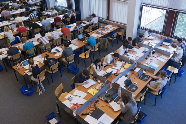 Ein großer Raum in einer Bibliothek, in dem viele Menschen an Schreibtischen mit Laptops Unterlagen arbeiten.