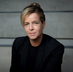 Portraitfoto einer Frau mit kurzem, blond-braunem Haar, schwarzem Oberteil und schwarzem Blazer vor grauem Hintergrund.