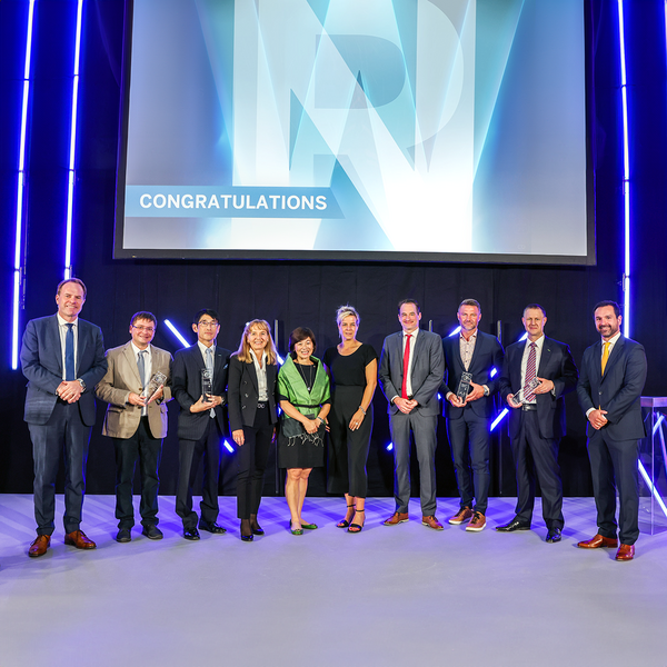 Ten people in business attire stand in a group, several holding glass trophies, with the word “Congratulations” displayed on a large monitor behind them.
