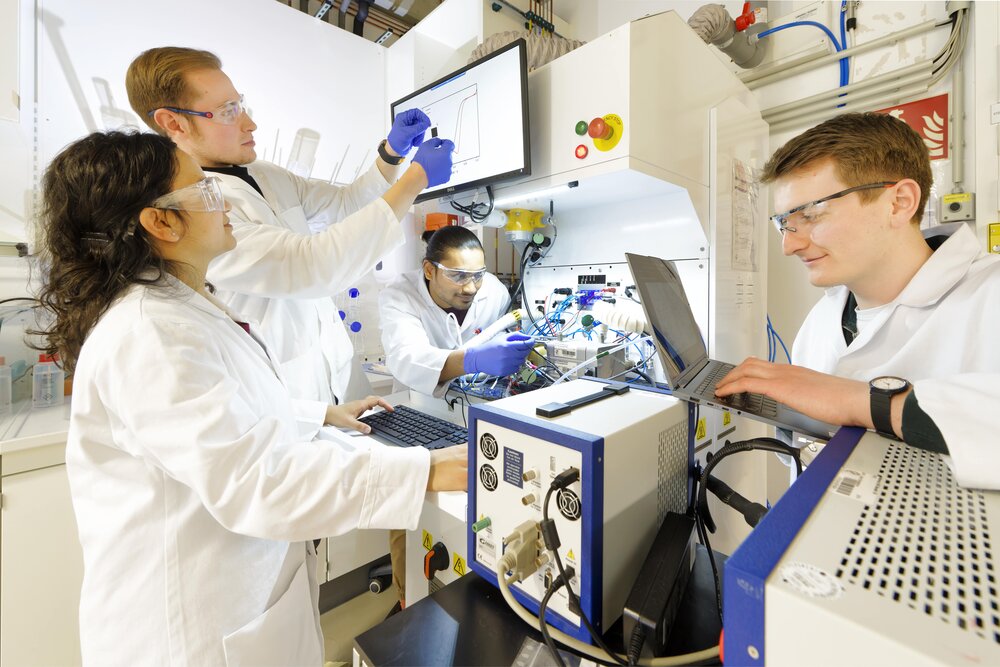 Four people wearing safety goggles and lab coats are working together on an experimental setup and looking at the screens of a laptop and monitor.