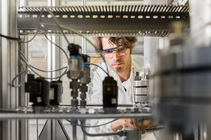 Ein Mann mit Schutzbrille und im Laborkittel arbeitet konzentriert an einer technischen Anlage mit Rohren und Ventilen.