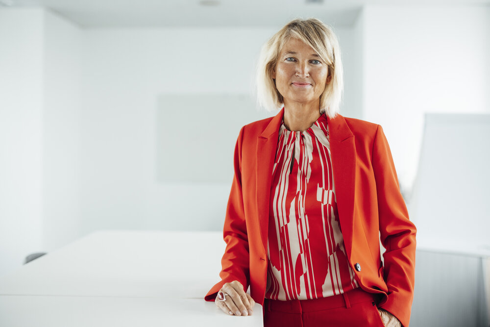 Ein Portraitfoto von einer  schick gekleideten Frau, die verschiedene orangefarbene Kleidungstücke trägt. Sie steht neben einem Bürotisch und setzt ihren Arm darauf ab.