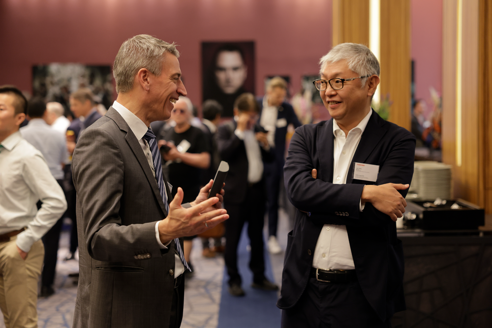 Two men in business attire engage in animated conversation at a networking event. The background is filled with other attendees and framed photos.