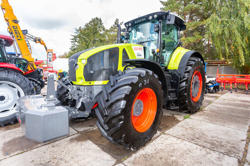 Ein grüner Traktor mit großen schwarzen Reifen und orangenen Felgen auf einer Fläche mit weiteren landwirtschaftlichen Geräten.