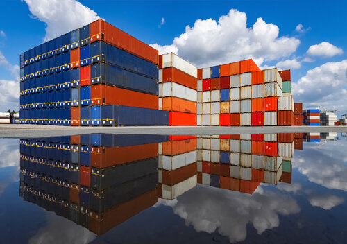 Gestapelte blaue, rote und weiße Container spiegeln sich in einer Wasserfläche unter blauem Himmel mit Wolken.