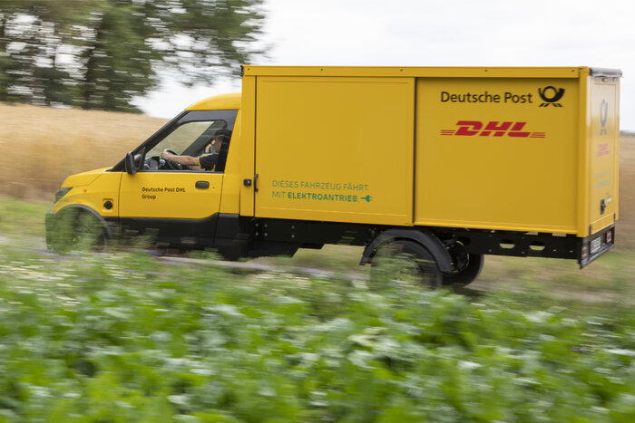 Ein gelber Lieferwagen der Deutschen Post DHL mit Elektroantrieb fährt durch ländliche Umgebung.