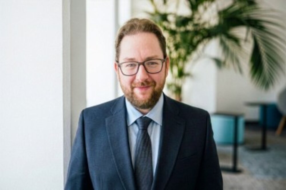 Man with glasses, wearing a suit and tie standing indoors with plants and furniture in the background