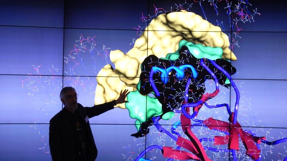 A man in a high- e presents a 3D model of molecular structures with colourful graphic elements on a large screen.