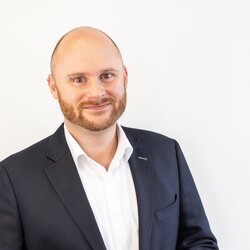 Ein Portraitfoto von einem Mann in einem Sakko über einem weißem Hemd, lächelnd vor weißem Hintergrund.