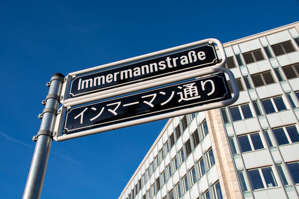 Ein Straßenschild mit der Aufschrift ‚Immermannstraße‘ in Deutsch und Japanisch, vor einem Bürogebäude.