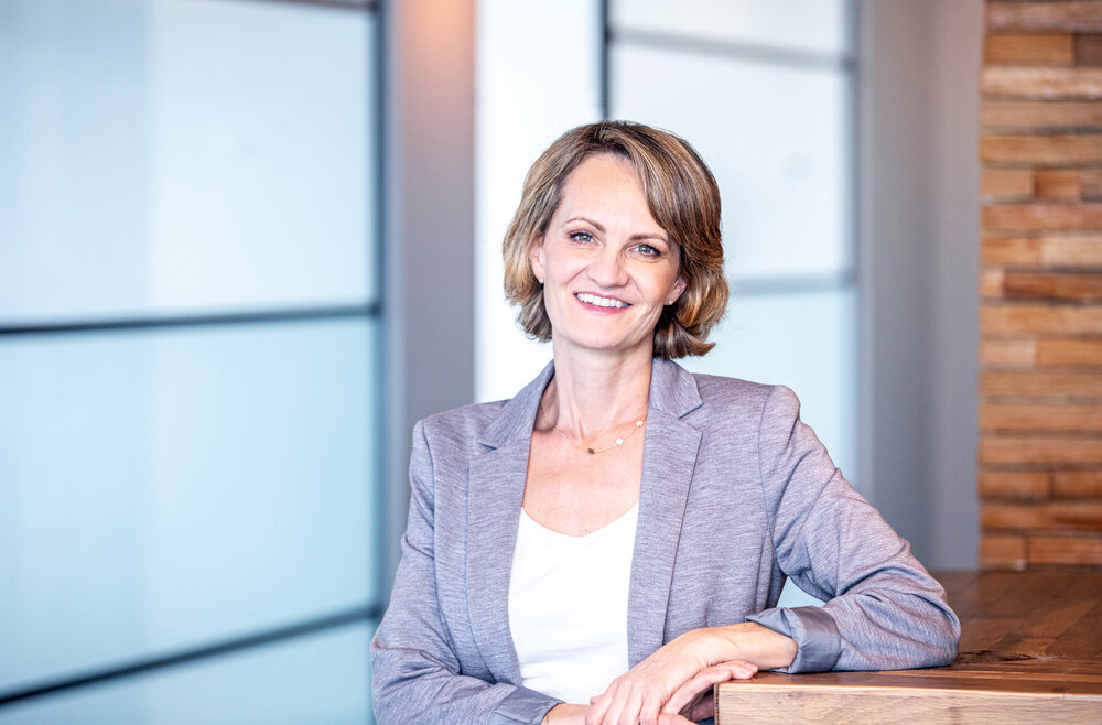 A portrait photograph of a smartly dressed woman with short light brown hair wearing a light gray blazer and white top sitting with hands resting on a wooden surface in a modern office setting