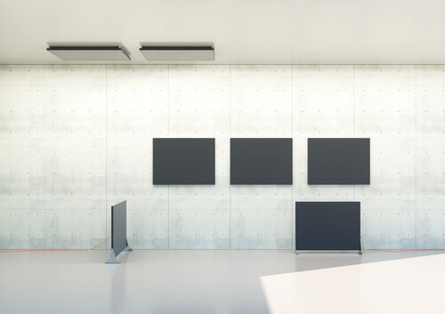 Ein minimalistischer Raum mit einer Betonwand, an der vier schwarze rechteckige Paneele hängen, zwei weitere stehen senkrecht auf dem Boden.