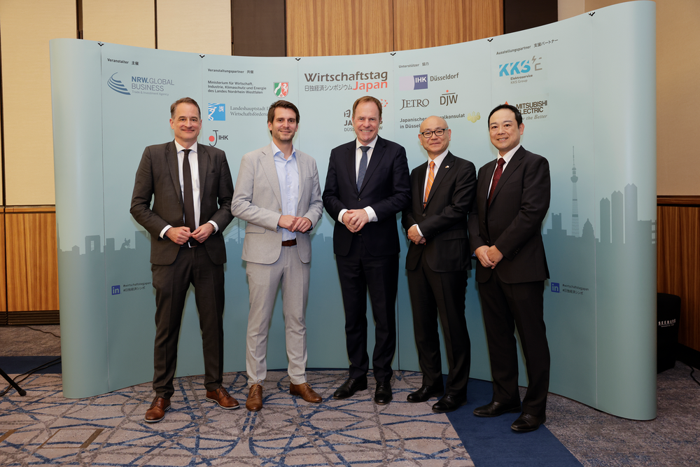 Five men in suits pose confidently in front of a backdrop with logos for a Wirtschaftstag Japan event, conveying professionalism and international collaboration.
