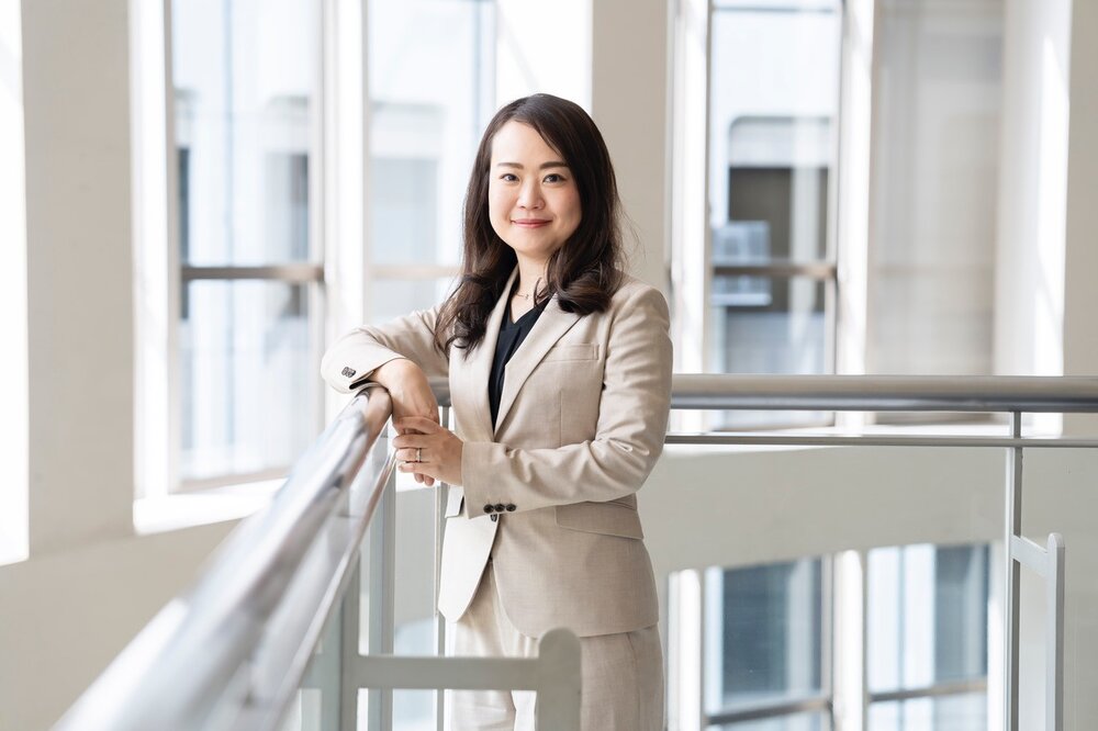 A portrait photograph of a smartly dressed woman in a light suit stands by a railing with one hand resting on it, smiling at the camera.