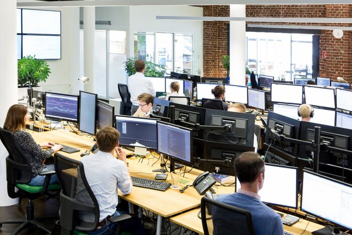 Großraumbüro mit mehreren Personen an Schreibtischen, umgeben von vielen Monitoren.