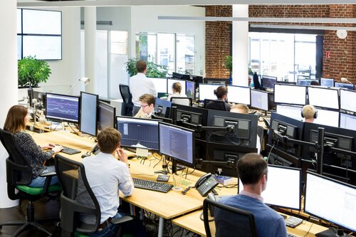 Großraumbüro mit mehreren Personen an Schreibtischen, umgeben von vielen Monitoren.