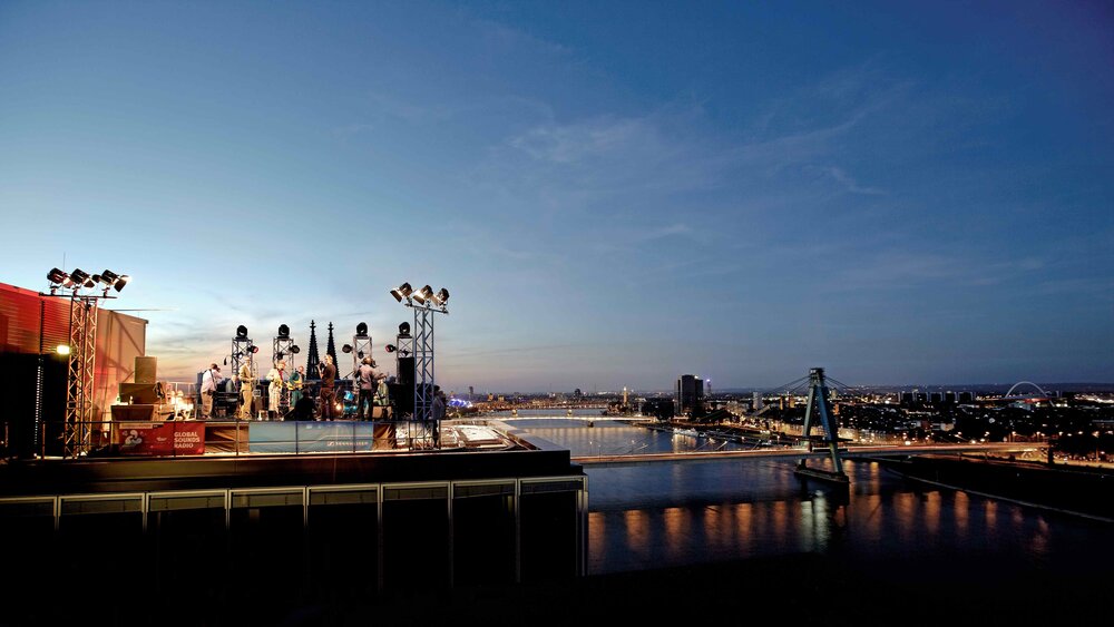 An open-air concert on a rooftop at sunset, with a view over a city and a river with illuminated bridges.