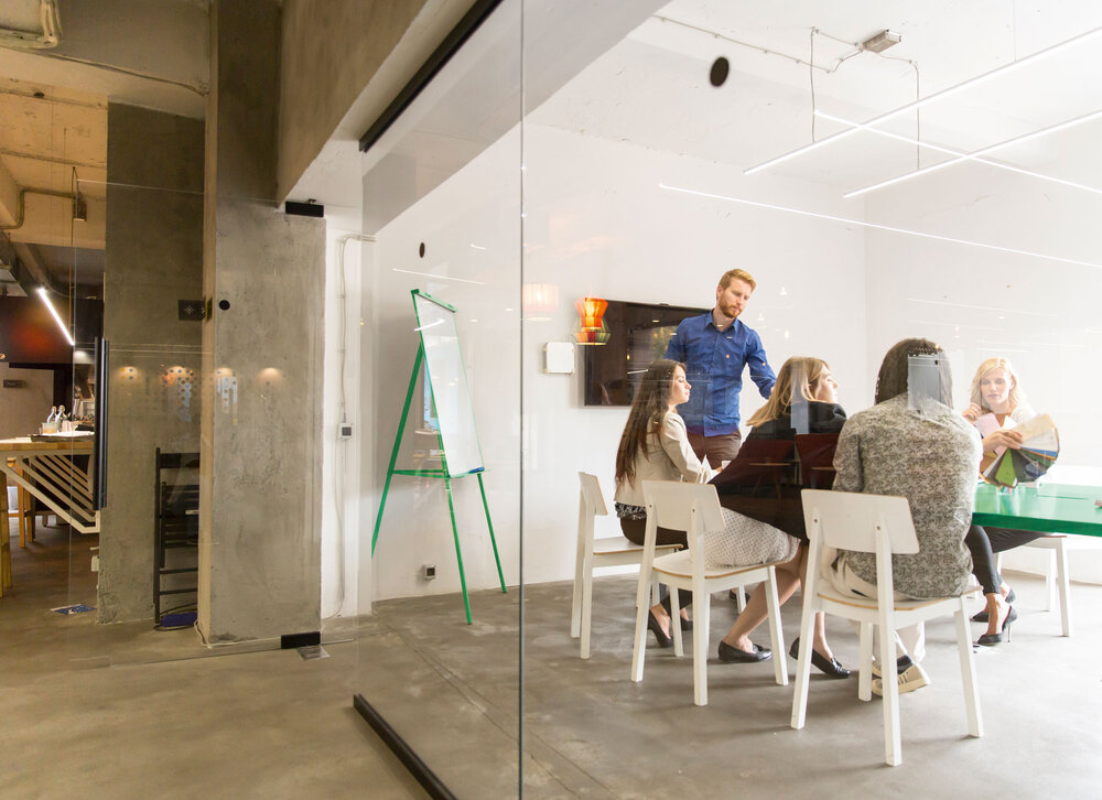 Several people in a modern conference room, sitting around a table, with a flip chart and glass walls nearby.
