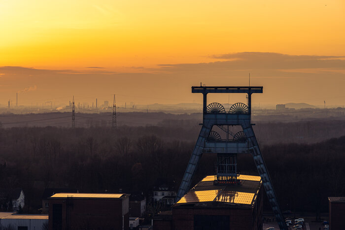 Der Förderturm einer ehemaligen Zeche im Vordergrund, dahinter Industrie- und Landschaftspanorama bei Sonnenuntergang.