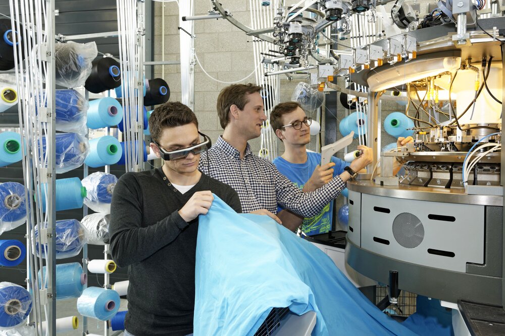 Three people are working at a textile machine, with blue spools of thread in the background and a large piece of blue fabric in the foreground.