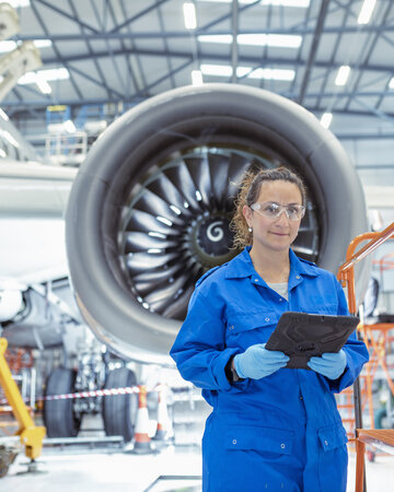 A woman in blue coveralls and safety glasses works on a tablet near a jet engine in a hangar, conveying focus and professionalism.