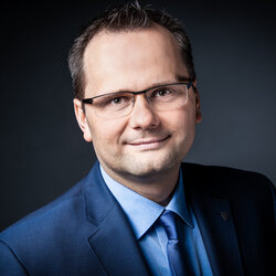 Porträt eines Mannes mit Brille im blauen Anzug, Hemd und Krawatte vor dunklem Hintergrund.