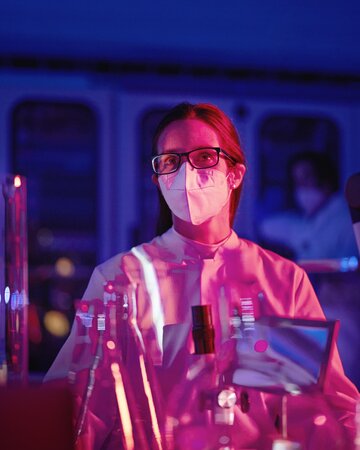 Eine Person mit Schutzmaske und Brille steht im Labor, beleuchtet von blauem und rotem Licht, vor ihr viele Reagenzgläser.