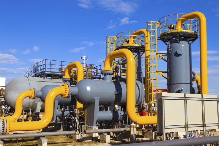Eine Industrieanlage mit gelben Rohrleitungen, grauen Druckbehältern und Leitern vor blauem Himmel.