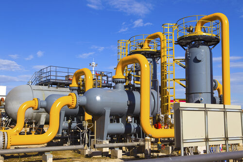 Eine Industrieanlage mit gelben Rohrleitungen, grauen Druckbehältern und Leitern vor blauem Himmel.